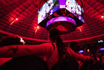 Photograph of a TCU women’s basketball player seen from a low rear angle, sitting with arms resting on folding chairs on either side during pregame introductions. A purple-lit jumbotron hangs overhead in a red-lit arena.