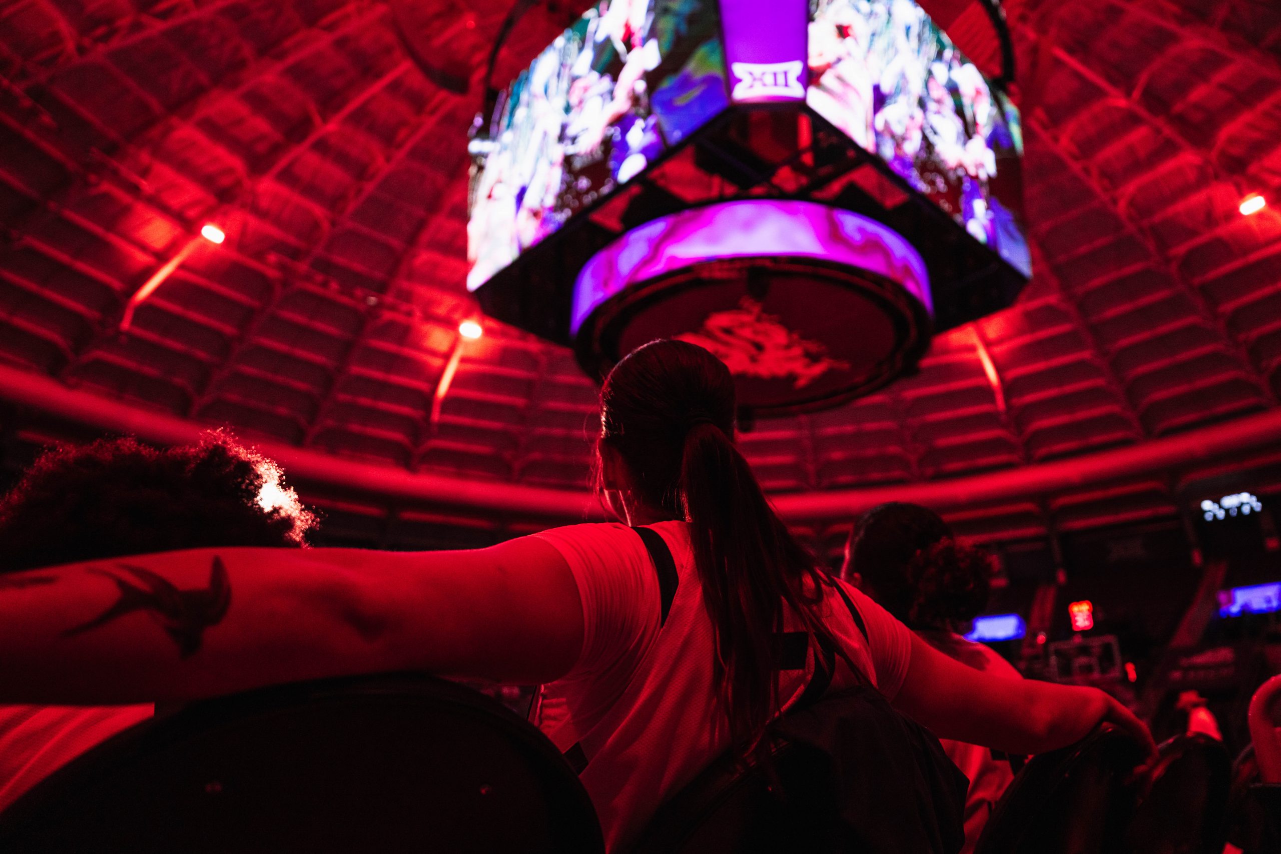Photograph of a TCU women’s basketball player seen from a low rear angle, sitting with arms resting on folding chairs on either side during pregame introductions. A purple-lit jumbotron hangs overhead in a red-lit arena.