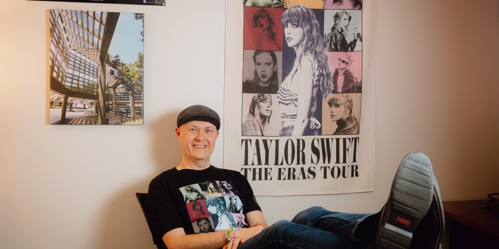Photograph of TCU professor and researcher Andrew Ledbetter seated in a black desk chair, smiling toward the camera with his feet up. He wears a black Taylor Swift “The Eras Tour” T-shirt, and a matching tour poster hangs on the wall behind him.