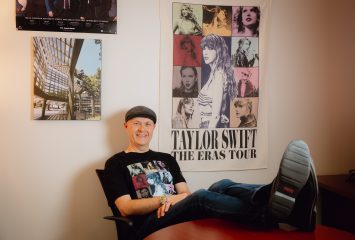 Photograph of TCU professor and researcher Andrew Ledbetter seated in a black desk chair, smiling toward the camera with his feet up. He wears a black Taylor Swift “The Eras Tour” T-shirt, and a matching tour poster hangs on the wall behind him.