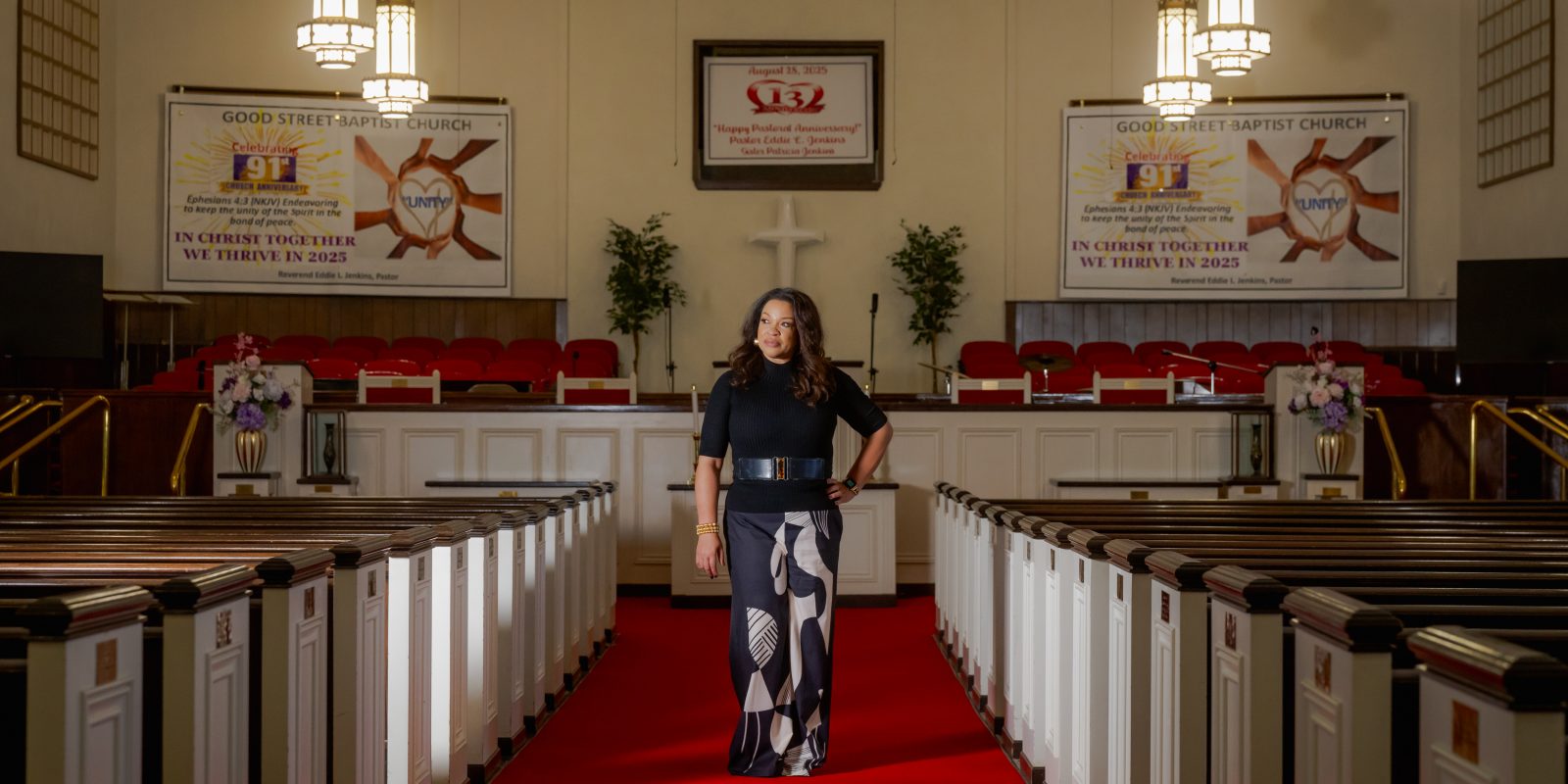 Photograph of TCU alumna L. Michelle Smith standing in a red-carpeted church aisle, flanked by white-sided wooden pews. Overhead lights hang above, with a tan wall in the background.