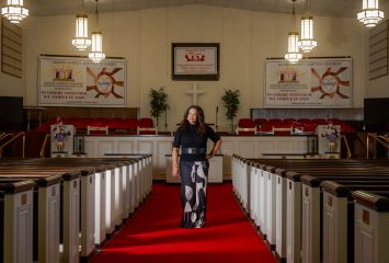 Photograph of TCU alumna L. Michelle Smith standing in a red-carpeted church aisle, flanked by white-sided wooden pews. Overhead lights hang above, with a tan wall in the background.