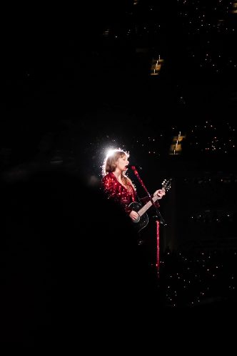Photograph of Taylor Swift performing in a dark setting, with her face, red sparkly garment, and red mic stand illuminated. A spotlight behind her is partially obscured by her head. Small yellowish lights speckle the right side of the mostly black background.