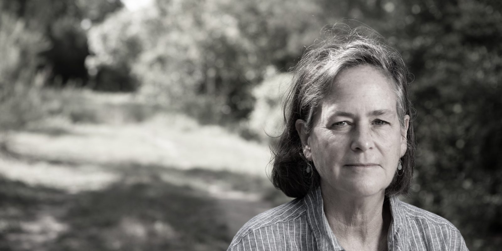 Black-and-white photograph of author Catherine O’Connor looking toward the camera, with a shadowy wood backdrop behind her.