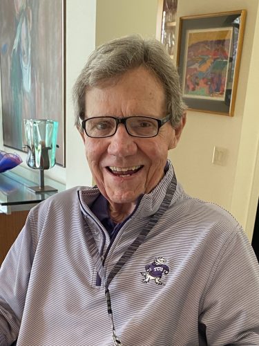 Photograph of TCU alumnus Buddy Dike wearing black glasses and a purple-and-white striped TCU zip-up, smiling at the camera in an indoor setting.