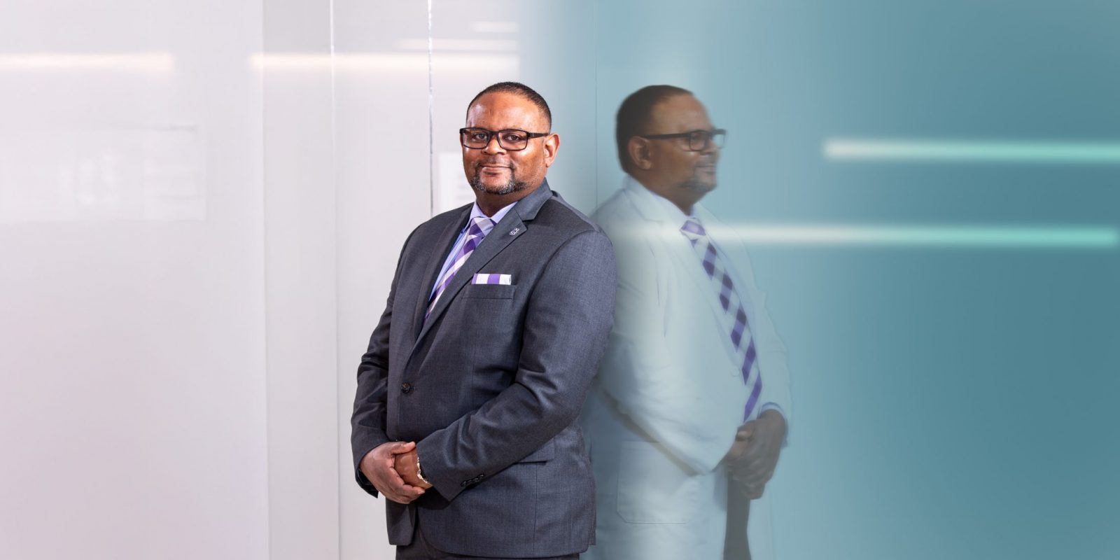 Photograph of TCU Provost Floyd L. Wormley Jr. standing with his hands clasped in front of him, wearing a dark grey business suit and tie. To his right, a profile reflection shows Wormley in a white coat, posed the same way.
