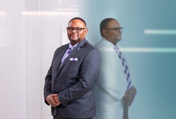 Photograph of TCU Provost Floyd L. Wormley Jr. standing with his hands clasped in front of him, wearing a dark grey business suit and tie. To his right, a profile reflection shows Wormley in a white coat, posed the same way.