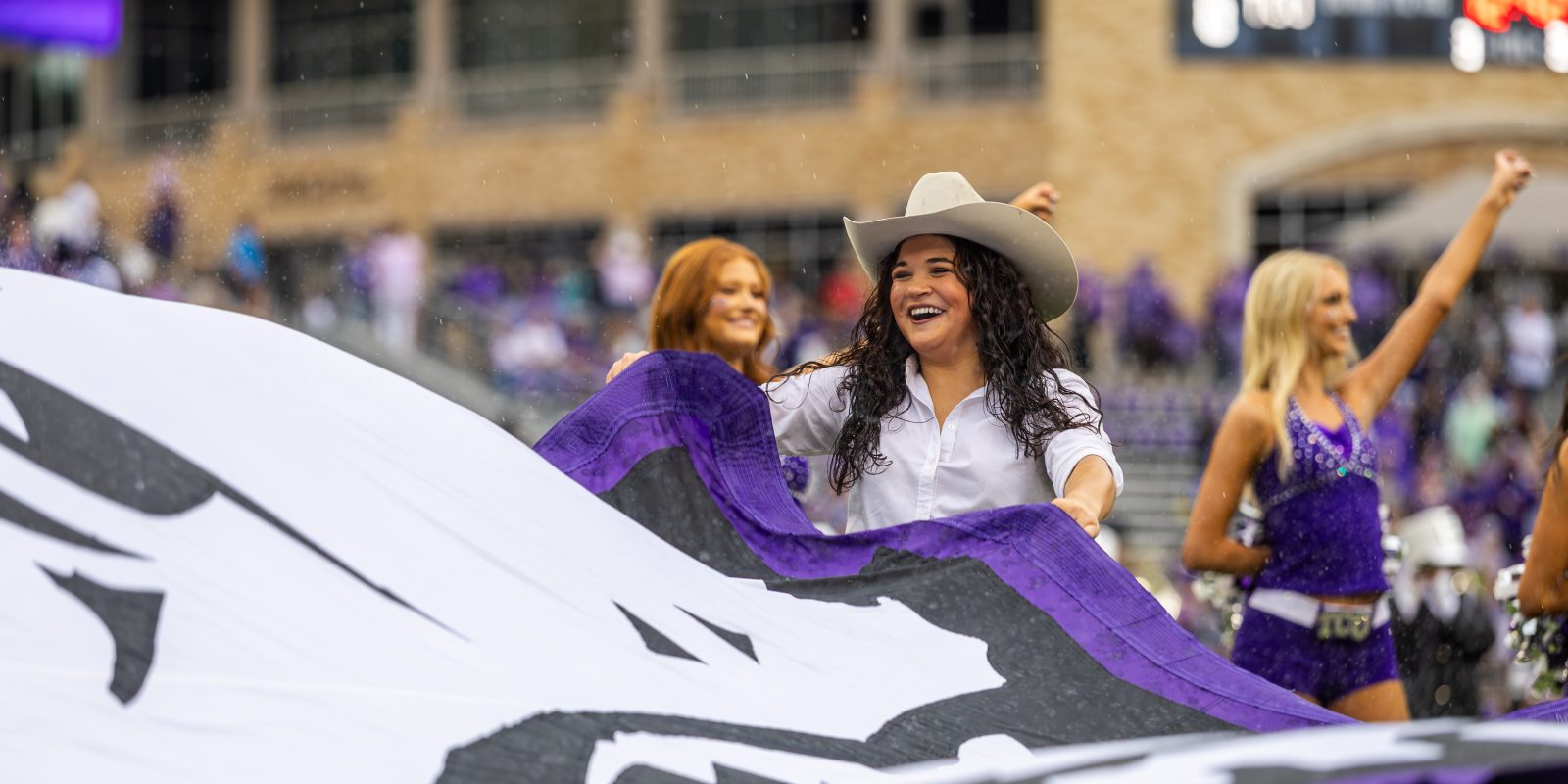 Image of a TCU Rangers spirit team member unfurling a giant purple TCU flag featuring a white and black horned frog on the field during a home football game. Cheerleaders and a large brick building are visible in the background.