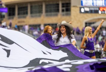 Image of a TCU Rangers spirit team member unfurling a giant purple TCU flag featuring a white and black horned frog on the field during a home football game. Cheerleaders and a large brick building are visible in the background.