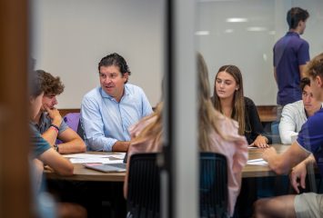 Photograph of Chancellor Daniel Pullin, in a professorial role, speaking to a group of students seated around a wooden table in a classroom. The image is framed by a window bar that divides the scene in half.