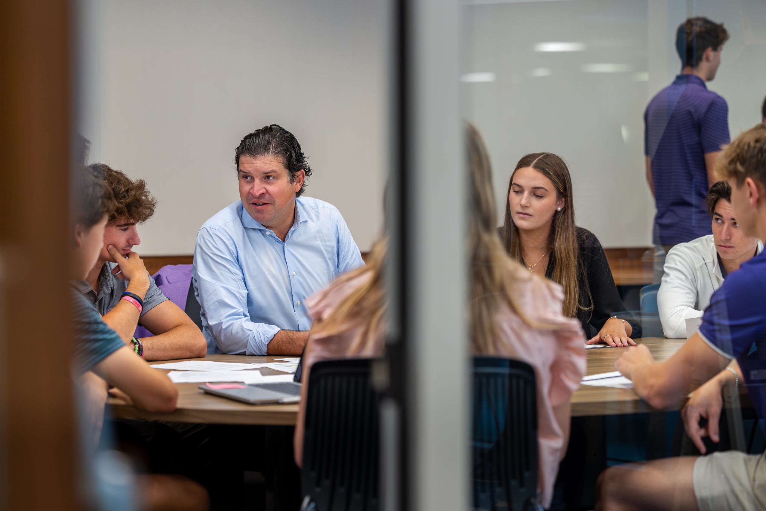 Photograph of Chancellor Daniel Pullin, in a professorial role, speaking to a group of students seated around a wooden table in a classroom. The image is framed by a window bar that divides the scene in half.
