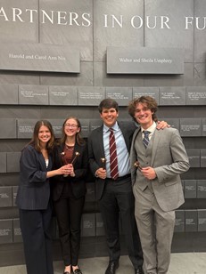 Photograph of four TCU Mock Trial team members in business attire, standing with their arms around each other’s shoulders and smiling at the camera against a grey stone wall.