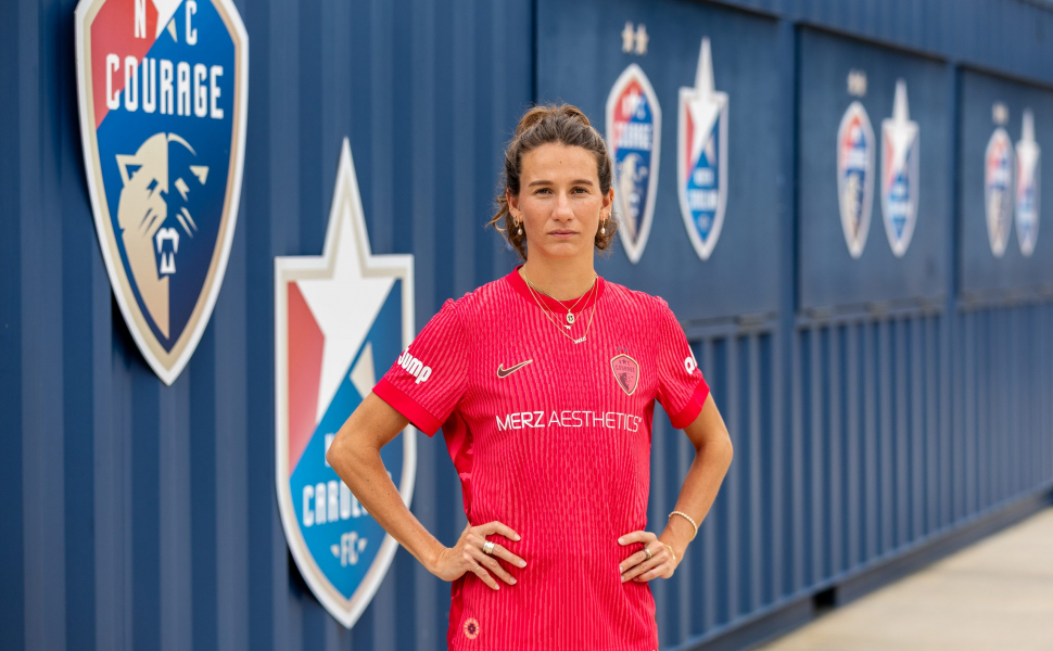 Photograph of TCU alumna and professional soccer player Ryan Williams standing with her hands on her hips in front of a navy blue wall decorated with North Carolina Courage emblems, wearing a red Courage uniform.