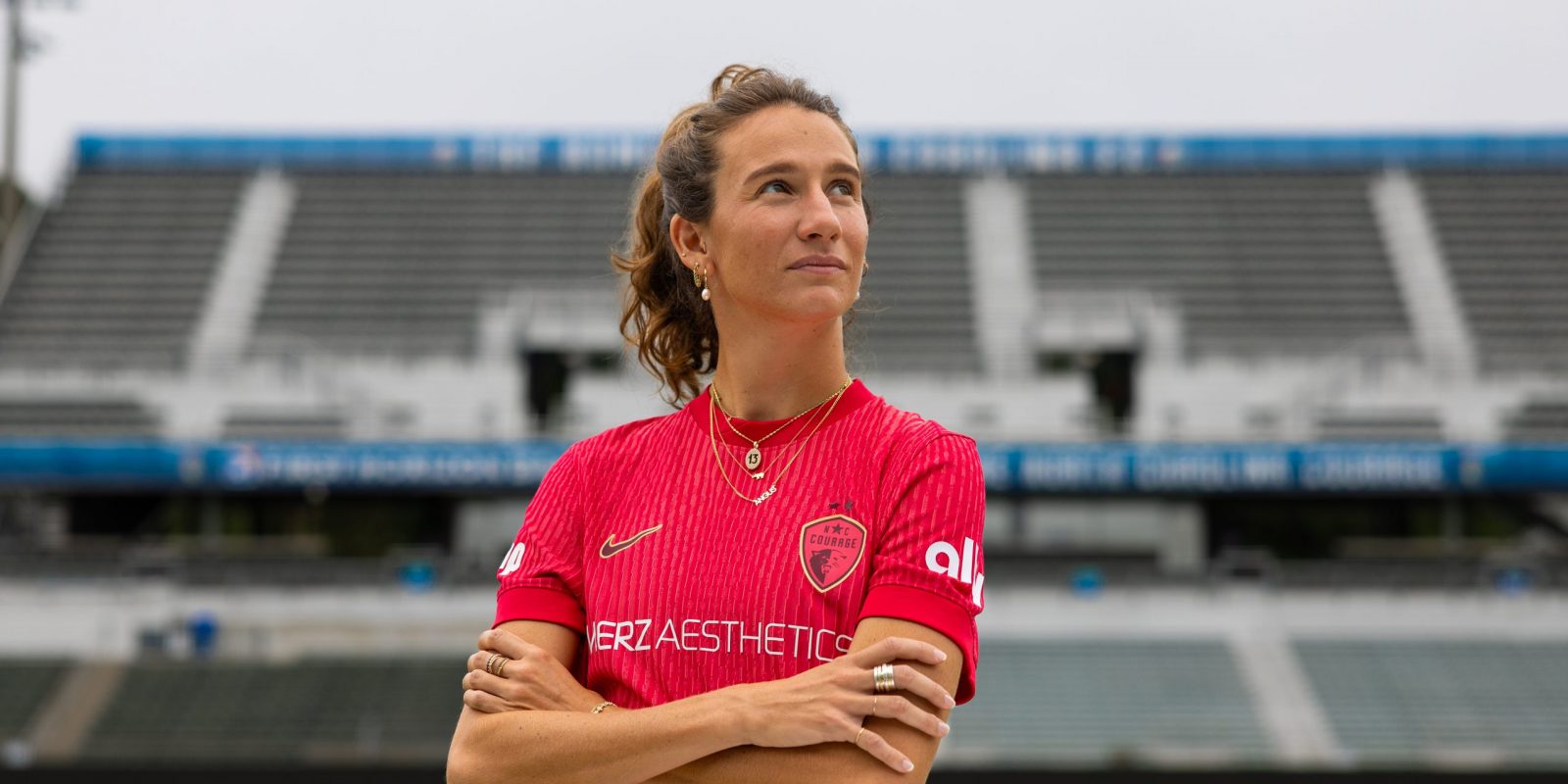 Photograph of TCU alumna and professional soccer player Ryan Williams in a red North Carolina Courage jersey, standing on a soccer pitch with her arms crossed and looking upward to her left.
