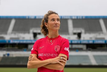 Photograph of TCU alumna and professional soccer player Ryan Williams in a red North Carolina Courage jersey, standing on a soccer pitch with her arms crossed and looking upward to her left.