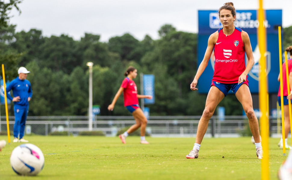 Photograph of TCU alumna and professional soccer player Ryan Williams, wearing a red practice jersey and blue shorts, watching a soccer ball in motion. Teammates, coaches and trees are visible in the background.
