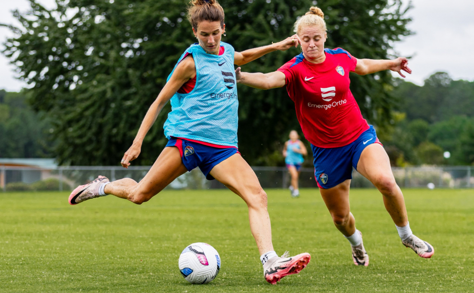 Photograph of TCU alumna and professional soccer player Ryan Williams, wearing a light blue practice pinnie and blue shorts, preparing to kick a mostly white soccer ball while a teammate in a red uniform and blue shorts chases from the side.