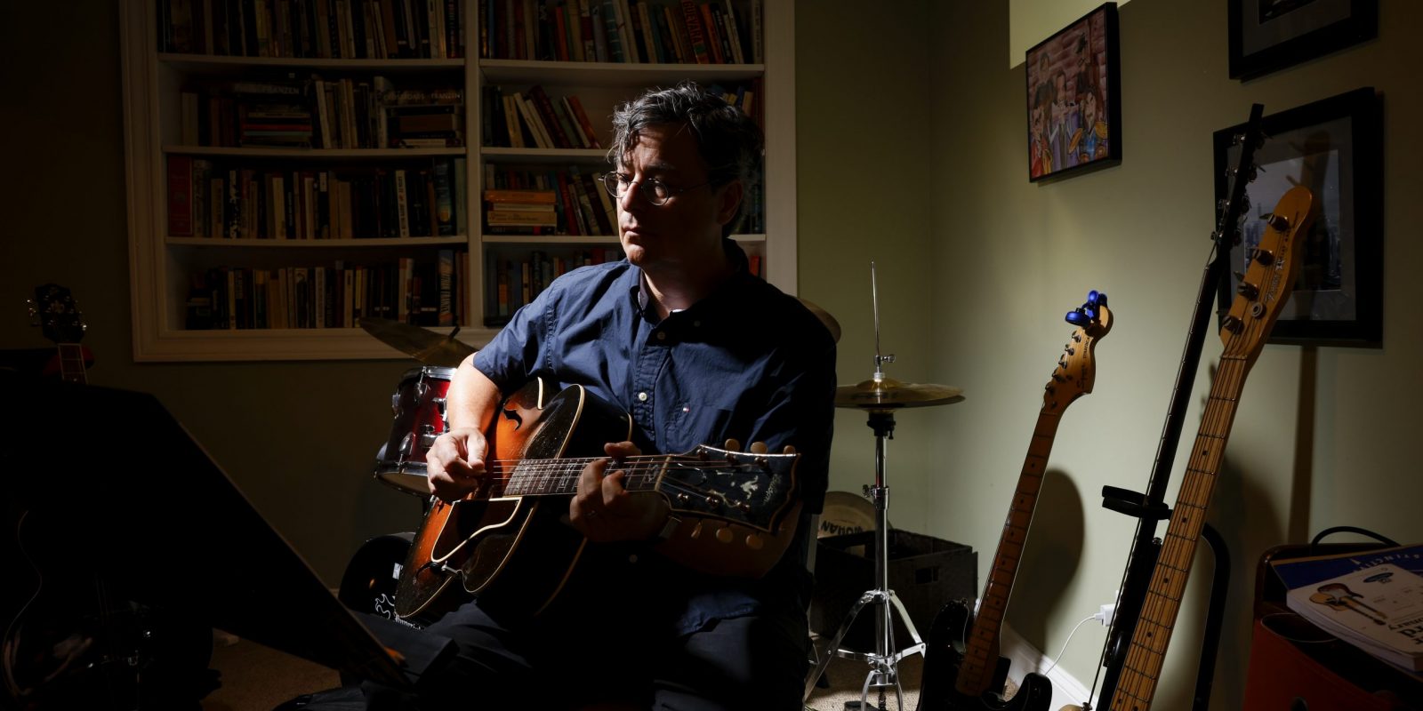 Photograph of TCU alum David Lay Williams seated in a dimly lit room, wearing a dark blue button-down shirt and holding a guitar across his lap.