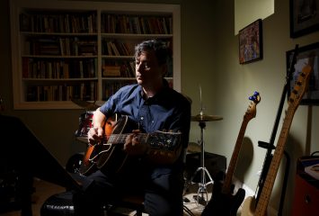 Photograph of TCU alum David Lay Williams seated in a dimly lit room, wearing a dark blue button-down shirt and holding a guitar across his lap.