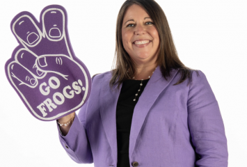 Studio photograph of TCU Dean of Admission Mandy Castro, wearing a purple sports jacket and black dress, holding a purple foam finger shaped like the "Go Frogs" hand sign with white text reading "Go Frogs" on the front.