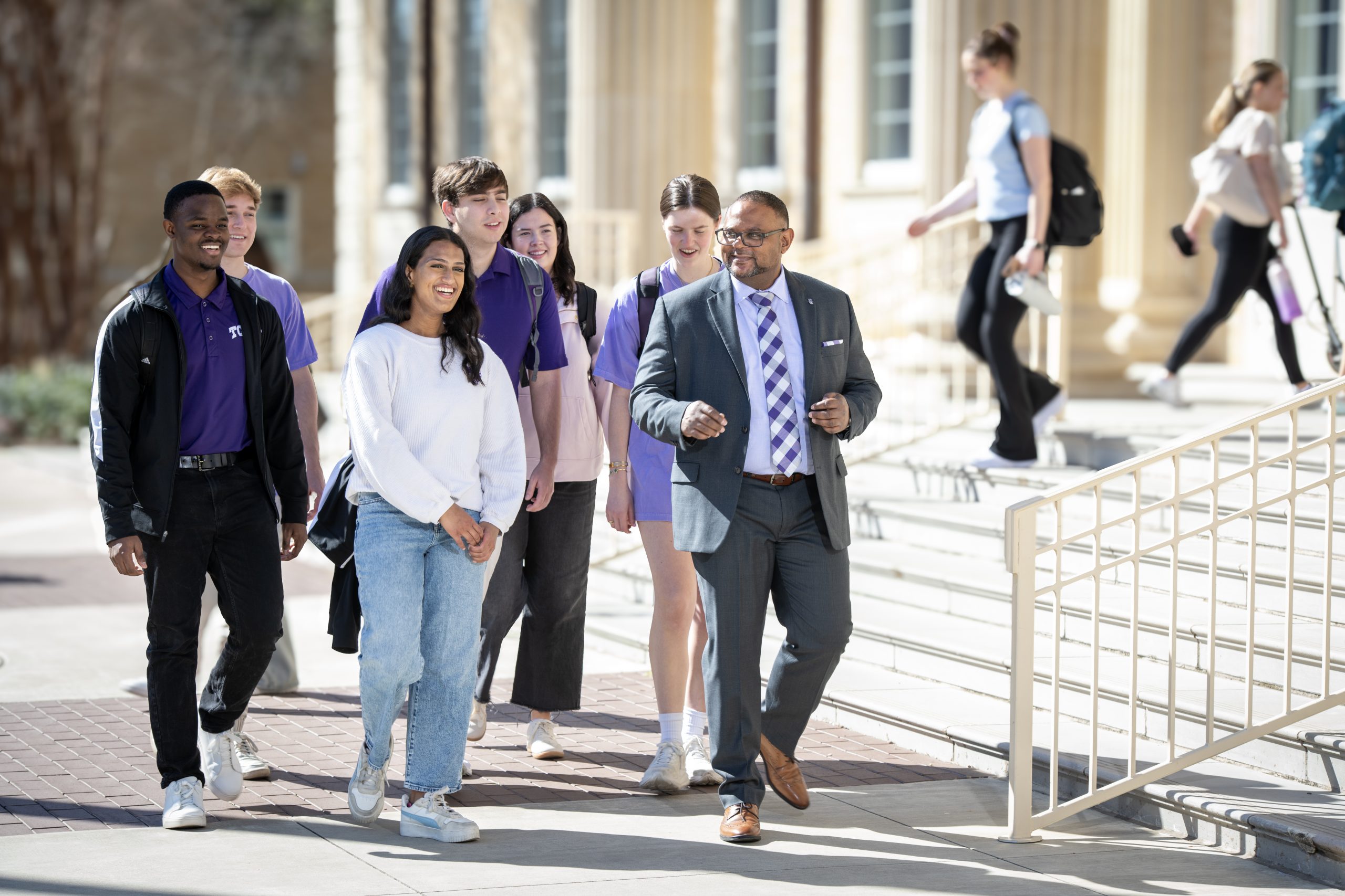 Photograph of TCU Provost Floyd L. Wormley Jr. in formal business attire walking across campus with six smiling students on a sunny day.