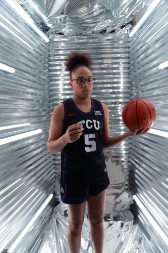 Photograph of TCU basketball player Olivia Miles in a black uniform holding a basketball in her left hand, standing against a well-lit silver metallic backdrop with horizontal ridges.