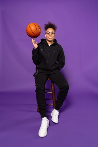 Photograph of Olivia Miles sitting on a stool against an all-purple backdrop, spinning a basketball on her right hand. She wears an all-black zip-up hoodie, black sweatpants and white sneakers, smiling toward the camera.