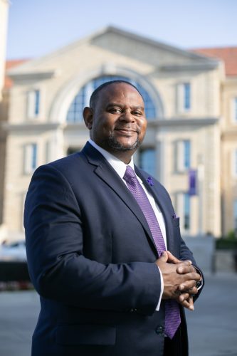 Photograph of TCU Provost Floyd Wormley Jr. smiling toothlessly at the camera, wearing a dark blue suit, white dress shirt and purple tie, with hands clasped over his stomach and a large campus building in the background on a sunny day.