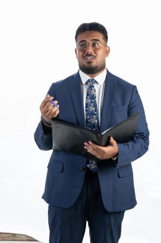 Photograph of TCU student Michael Shehata, dressed in a dark blue blazer, white button-down shirt and dark blue tie, holding a black folder in his left hand while gesturing toward the camera with his right.