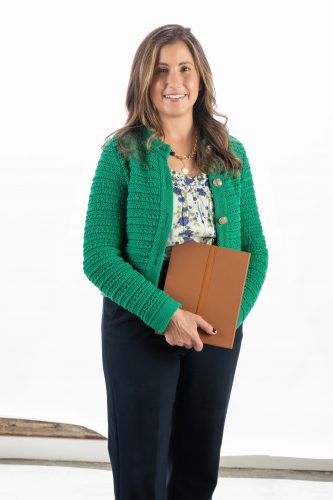 Studio photograph of Michele Meitl, associate professor of criminology and criminal justice, smiling at the camera while holding a brown notebook or file, dressed in professional attire.