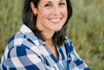 Photograph of TCU alumna Katie Becker seated on a grassy lawn, smiling at the camera while wearing a white-and-blue plaid shirt and blue jeans.