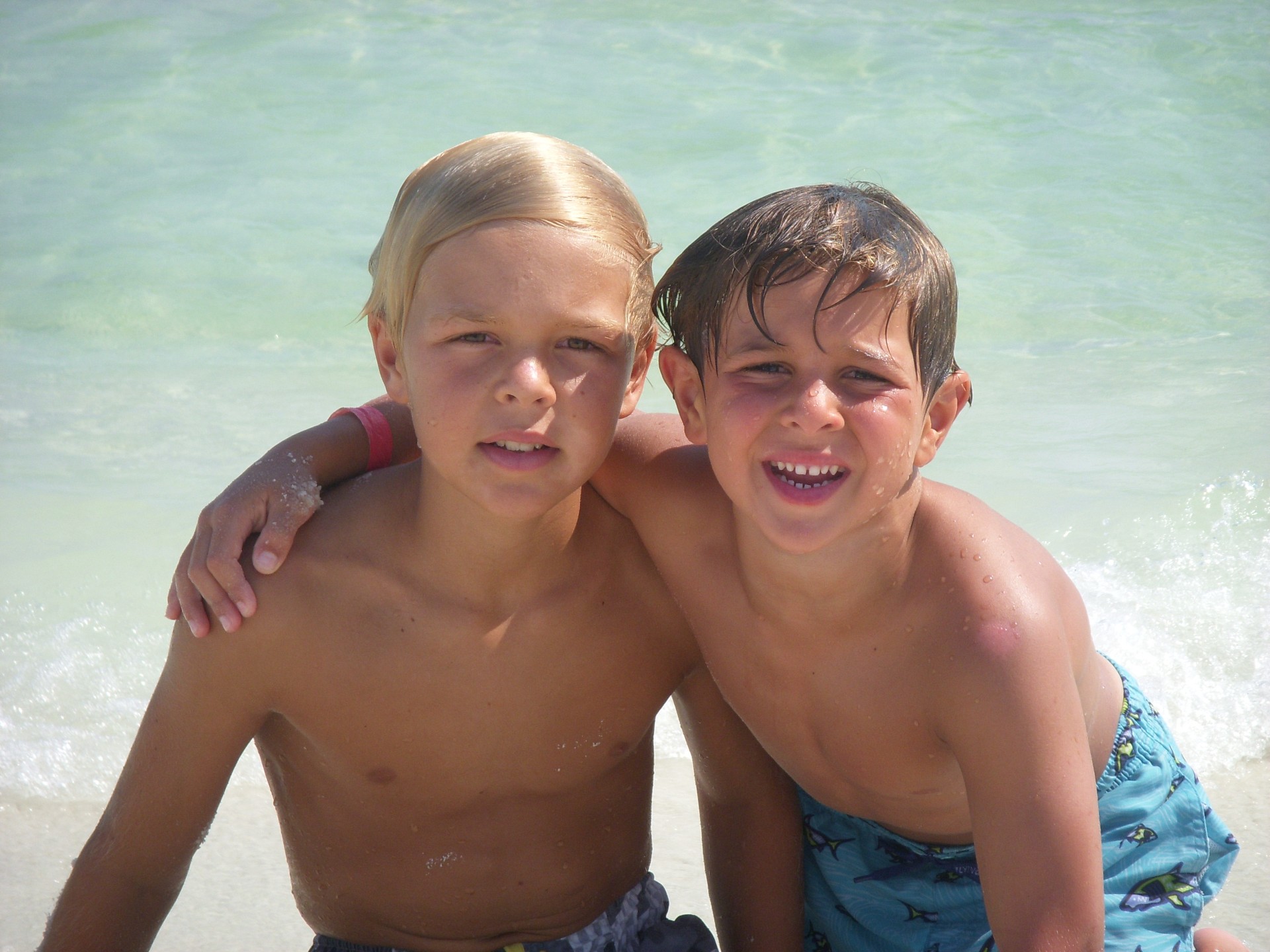 Photograph of the Bech brothers, Tiger on the left and Jack on the right, kneeling on sand with their arms around each other’s shoulders, wearing bathing suits and smiling.