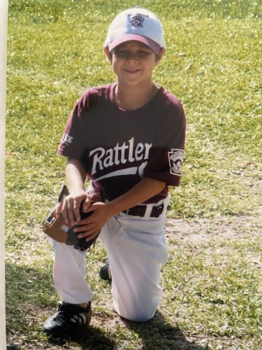 Photograph of Jack Bech as a young child dressed in a white and maroon Little League baseball uniform with the team name “Rattlers” in white font across his chest. Jack kneels with his hands and baseball glove resting on his right knee, squinting against the sunlight and smiling.