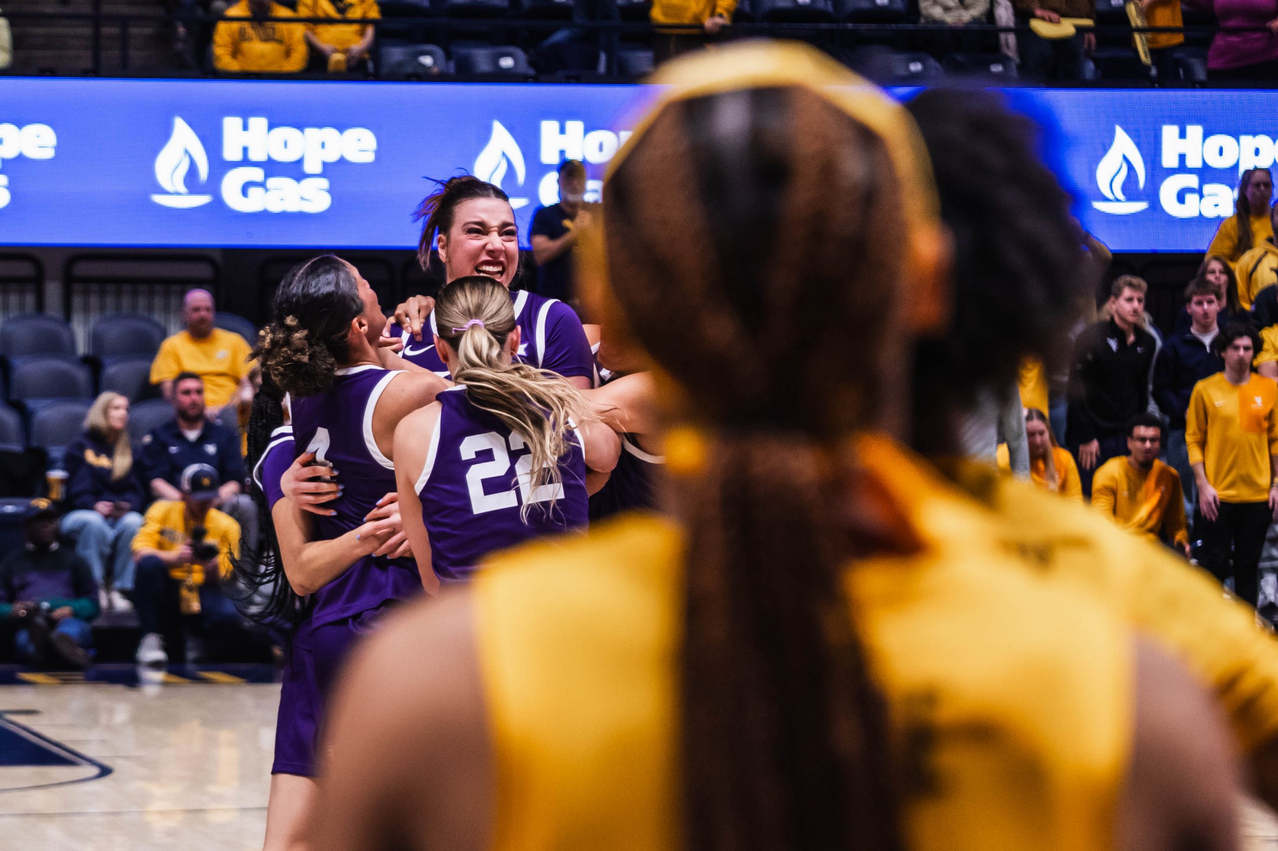 Photograph of TCU women’s basketball players celebrating with Marta Suárez after her game-winning shot against West Virginia in a January 2026 NCAA game. Two West Virginia players in yellow uniforms stand with their backs to the camera in the foreground.