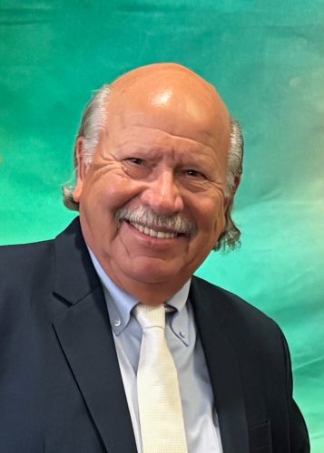 Portrait of longtime TCU administrator Don B. Mills, wearing a dark blue or black sport coat, light tan tie and dress shirt, as he smiles, photographed against an aqua green background.