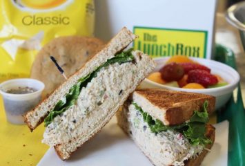 Photograph of a tuna and lettuce sandwich on toasted bread, sliced in half, with a fruit cup, a yellow bag of chips, a cookie and a white box labeled “The Lunch Box” in the background.