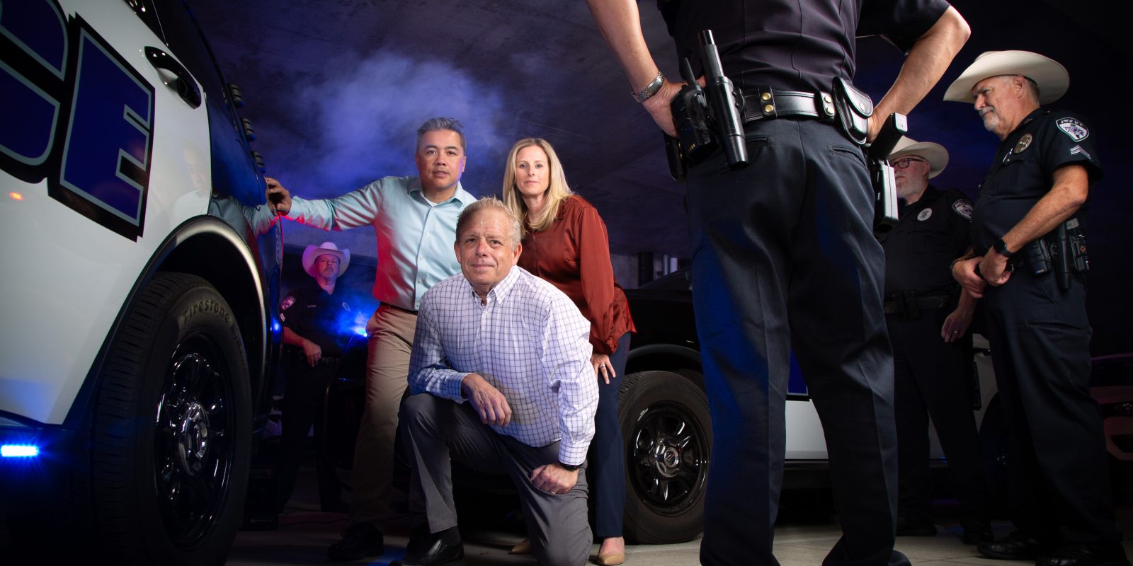 TCU criminal justice professors Johnny Nhan, Ronald Burns and Kendra Bowen stand with North Texas law enforcement officers near a police vehicle in a training facility.