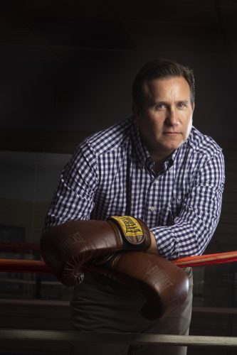 Photograph of Christopher Watts leaning against a red rope as he stands on the edge of a boxing ring wearing a checkered collared shirt and brown Everlast boxing gloves.