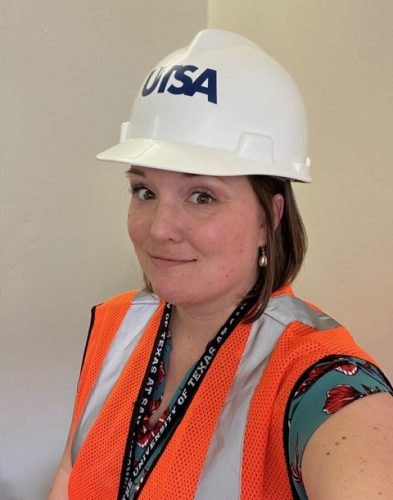 Stephanie Loken Edgett wears a white UTSA hard hat and orange safety vest on a construction site.