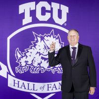 Richard Sybesma, TCU Swim & Dive coach and alumnus, displays the “Go Frogs” hand sign while standing in front of a purple TCU Hall of Fame banner.