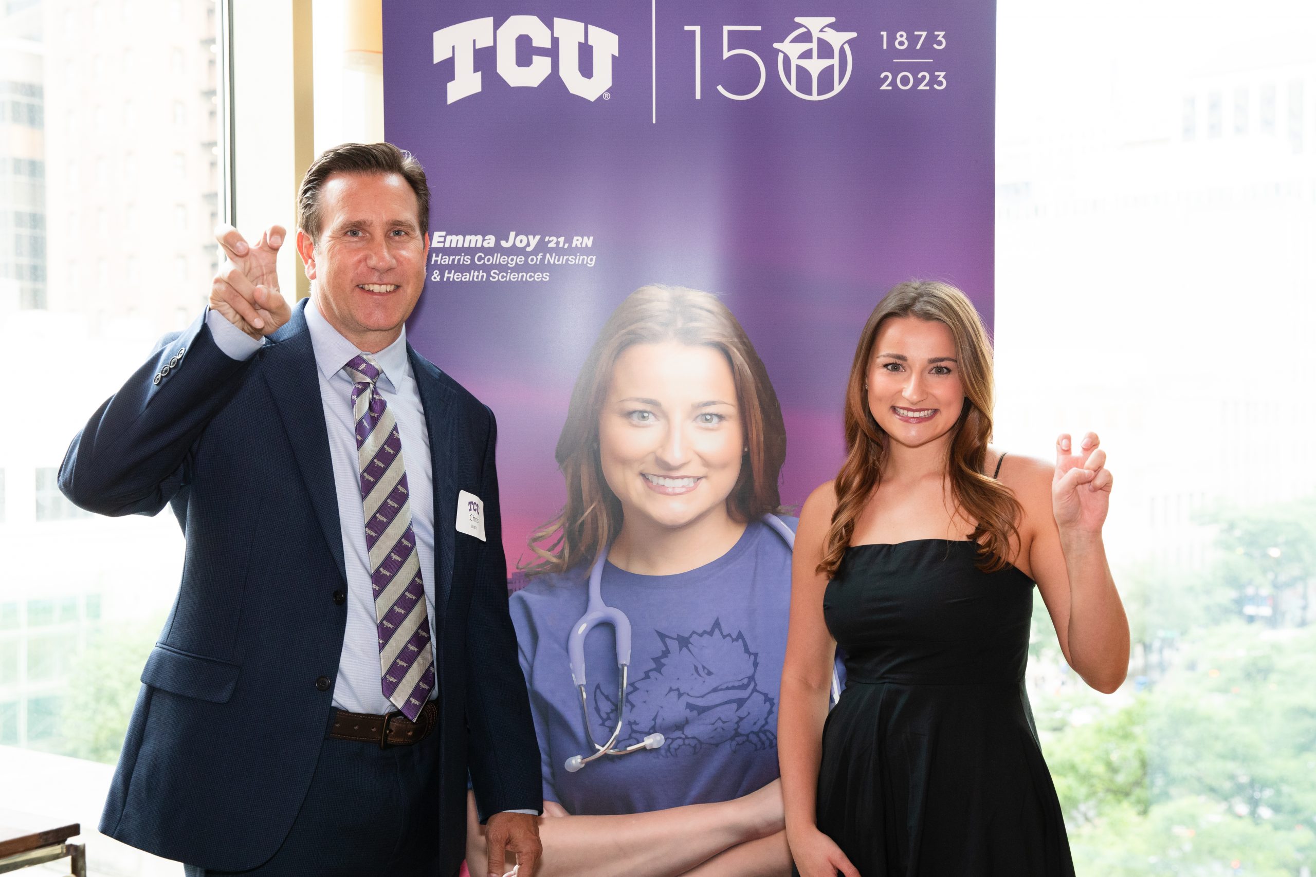 Photograph of Christopher Watts, wearing a suit, dress shirt and striped tie, standing beside a poster featuring a TCU student and the student herself, both making the “Go Frogs” hand sign as they smile.
