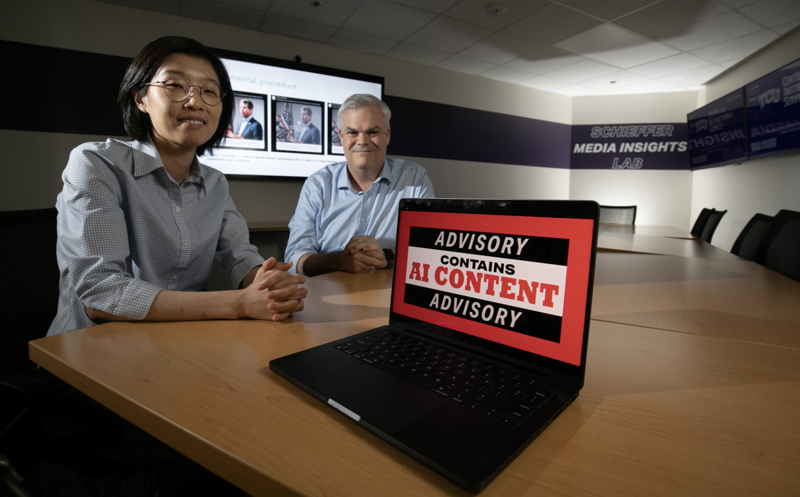 Photograph of TCU professors Jie Zhuang and Daxton “Chip” Stewart seated at a long wooden conference table in the Schieffer Media Insights Lab. They smile beside a laptop displaying a red warning screen reading “Advisory: Contains AI Content,” while a large monitor behind them shows images labeled “Experimental procedure.”