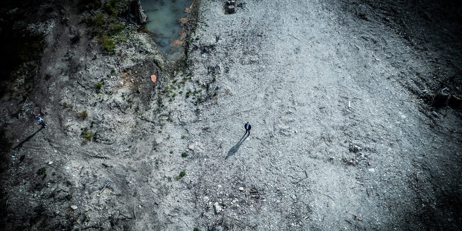 Aerial drone view of a lone figure standing in the vast, barren expanse of flood-damaged Louise Hays Park in Kerrville, Texas. The person casts a long shadow across the gray, debris-strewn ground. A small pool of murky water and sparse patches of vegetation are visible at the upper left.