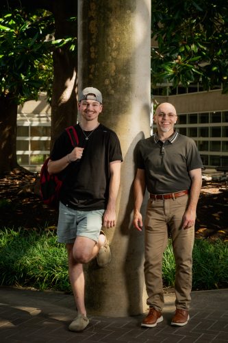 Paul Schrodt and his son, Philip, stand beside a tan stone pillar in a shaded area of TCU’s campus.