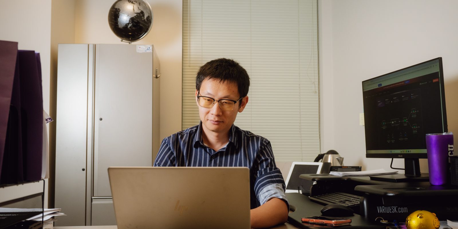 Xiaolu Zhou works on a laptop at his desk in his TCU office, with a globe visible on a shelf behind him.