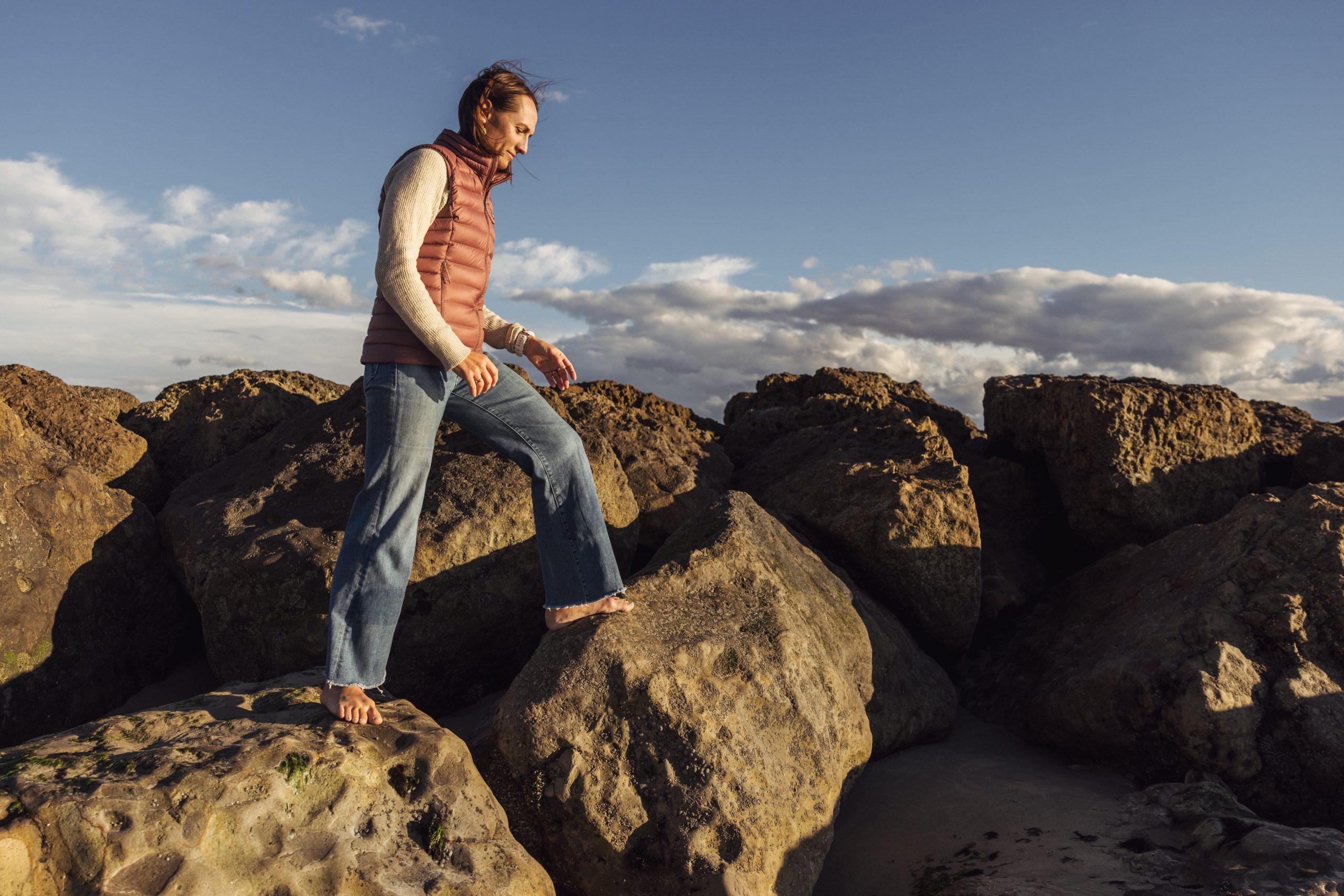 Kim Drenner walks barefoot on coastal rocks wearing a coral puffer vest, cream sweater and jeans, looking down against a backdrop of dramatic cloudy skies at golden hour.