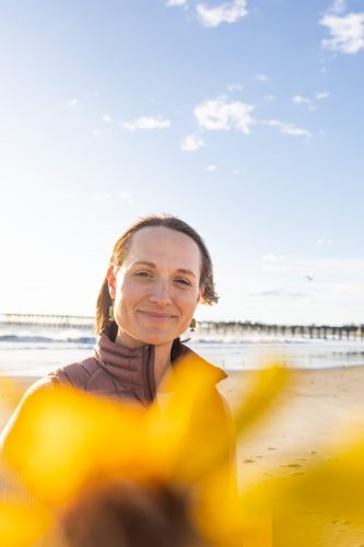 Kim Drenner smiles at the camera while standing on a beach at golden hour, wearing a brown puffer jacket over a yellow garment, with a wooden pier visible in the background.