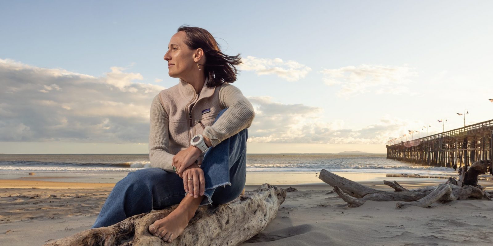 Kim Drenner, TCU alumna and head of environmental impact for Patagonia, sits barefoot on driftwood at a beach during golden hour, wearing a Patagonia sweater and jeans, with her hair blowing in the wind as she gazes thoughtfully toward the ocean. A wooden pier extends into the water in the background.