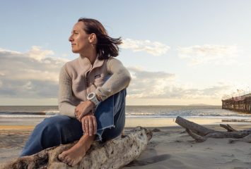 Kim Drenner, TCU alumna and head of environmental impact for Patagonia, sits barefoot on driftwood at a beach during golden hour, wearing a Patagonia sweater and jeans, with her hair blowing in the wind as she gazes thoughtfully toward the ocean. A wooden pier extends into the water in the background.