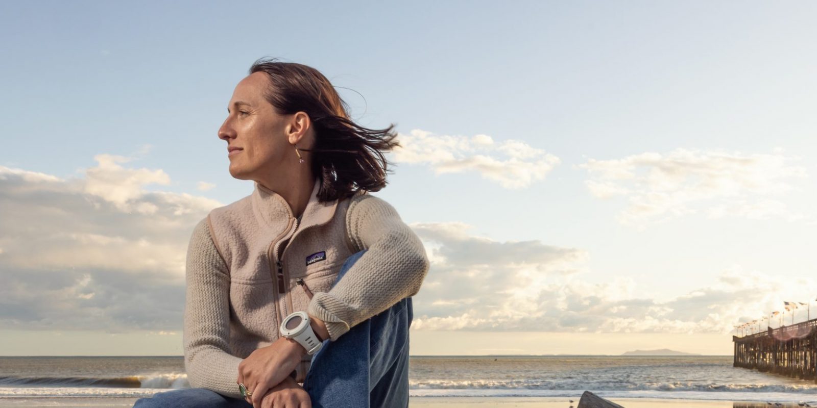 Kim Drenner, TCU alumna and head of environmental impact for Patagonia, sits barefoot on driftwood at a beach during golden hour, wearing a Patagonia sweater and jeans, with her hair blowing in the wind as she gazes thoughtfully toward the ocean. A wooden pier extends into the water in the background.